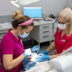 Dwie dentystki w maseczkach i rękawiczkach wykonują zabieg u pacjenta.